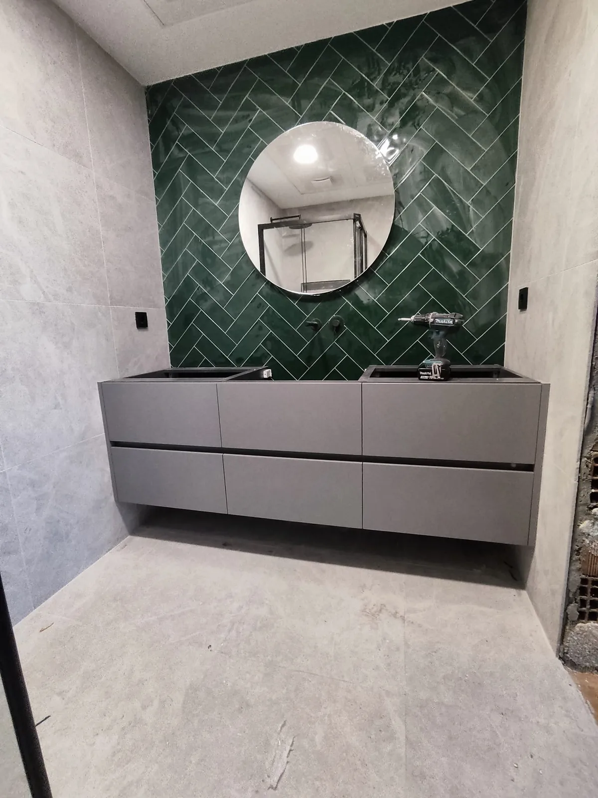 Yeşil duvar önünde geniş gri asma banyo dolabı ve yuvarlak ayna